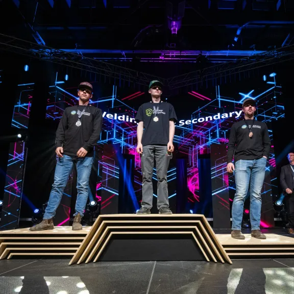 Lucas Stang (RDP Welding student) stands with his gold medal on a podium with two other welders at Skills Canada Alberta 2025