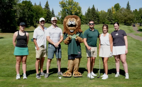 A group of golfers, student athletes, and Rufus the lion stand on a golf course fairway