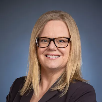 Laurel in a black blazer against a blue background smiles at the camera. She has shoulder length blonde hair and black rectangular glasses.