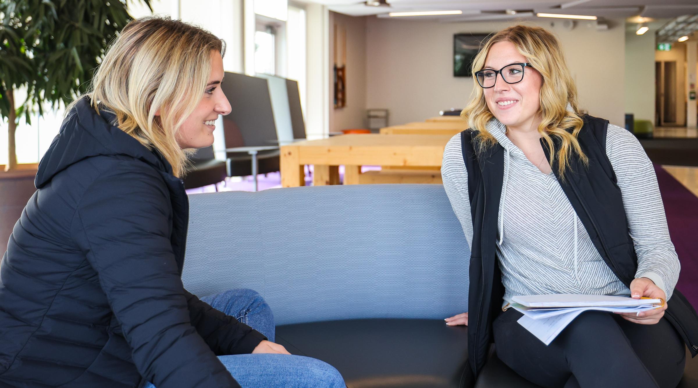 Two blonde women look at each other, smiling. One is holding a note book.