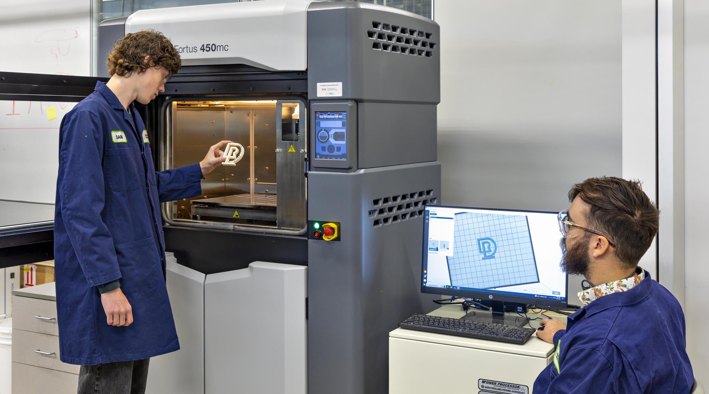 A student is pulling a 3D printed RDP logo out of a 3D printer while a faculty member watches