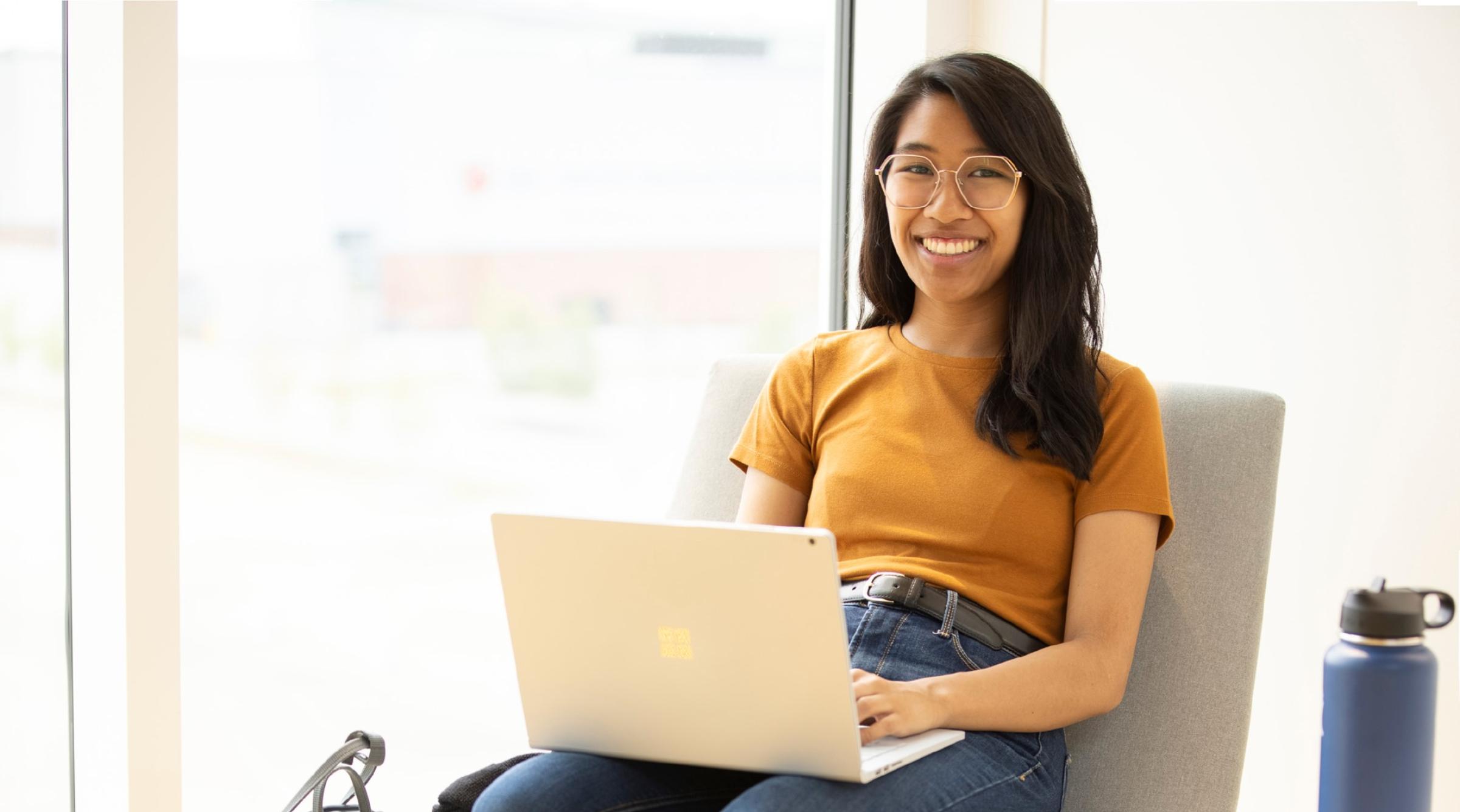 Smiling student sitting in a chair with a laptop on her lap in front of a bright window