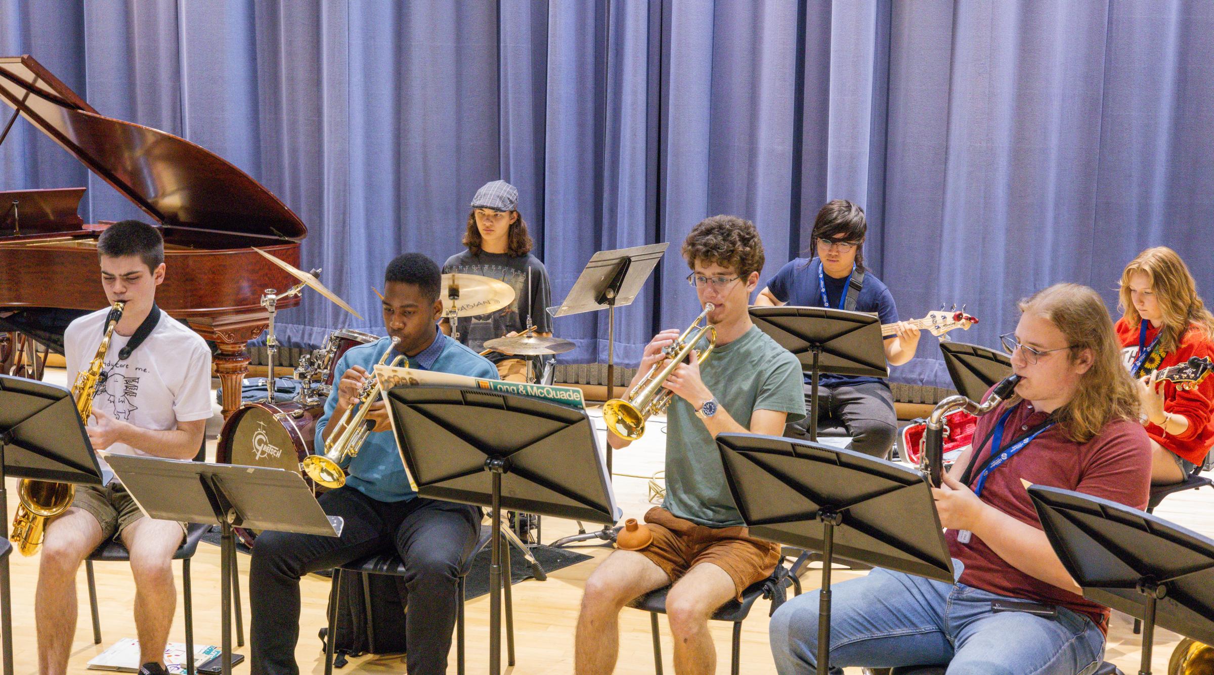 A diverse group of students sit in rows playing various instruments in front of music stands