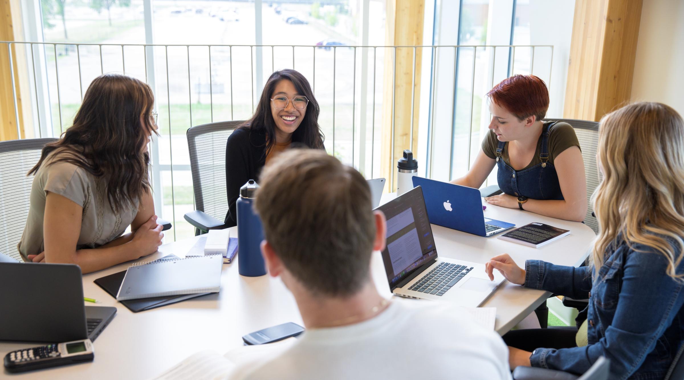 A group of smiling diverse students sits around a table with their laptops and books out. 