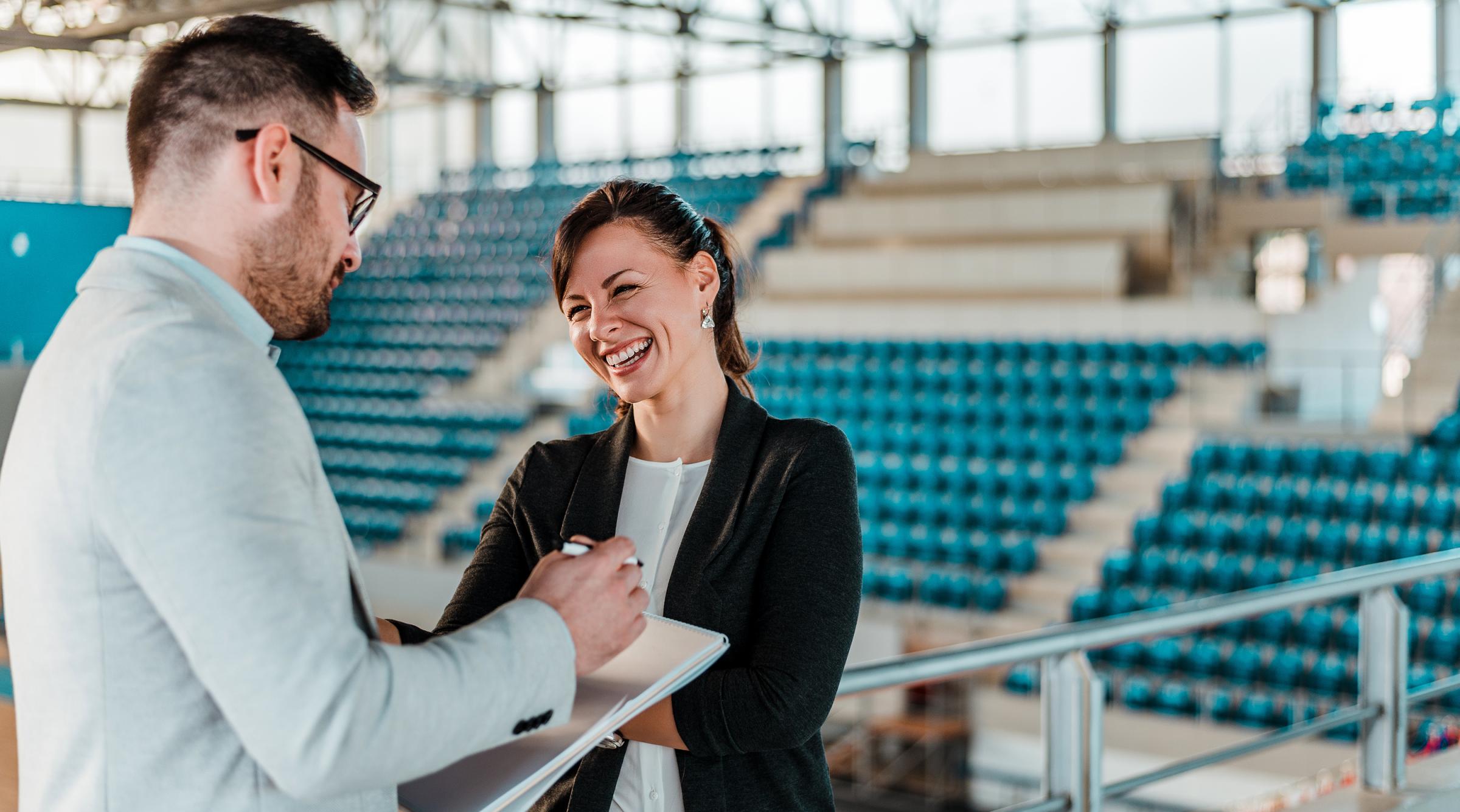 A man with a clipboard and a smiling woman stand in front of a large gymnasium