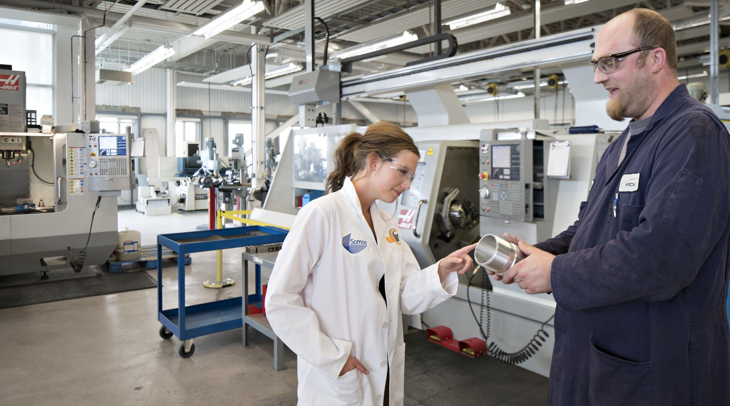 A female student in a lab coat examines a part being shown to her by a male lab tech in the CIM-TAC