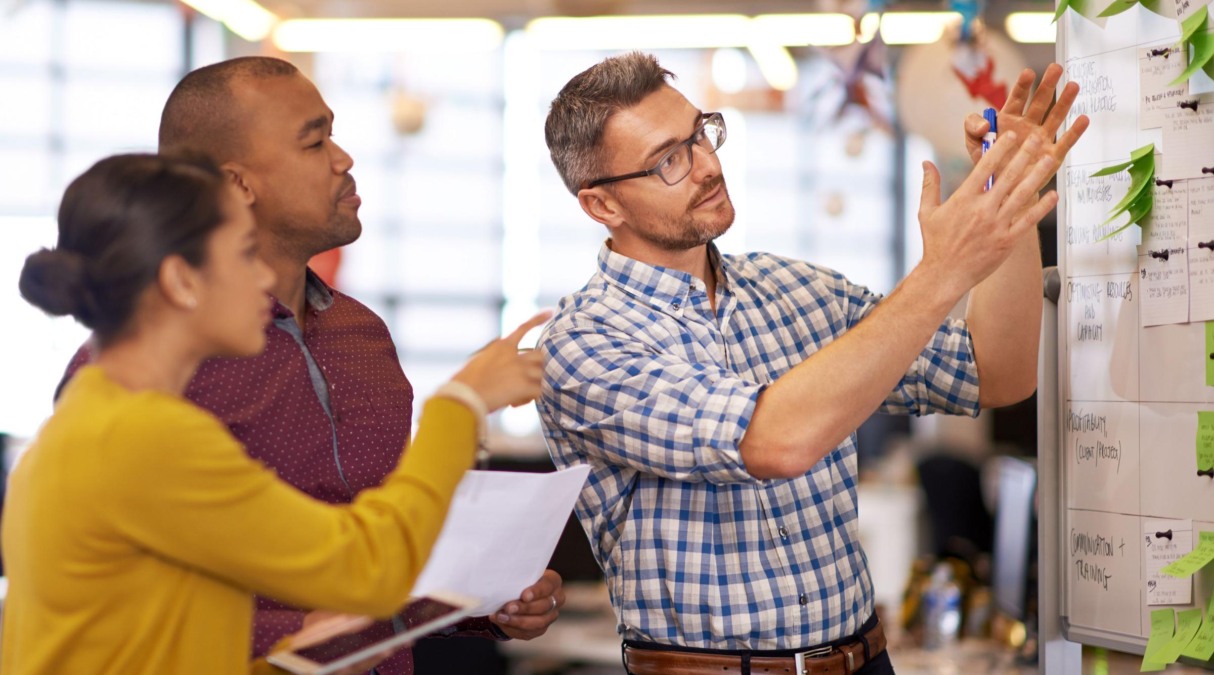 Two men and one woman in an office talk and point towards a whiteboard covered in writing and post-its