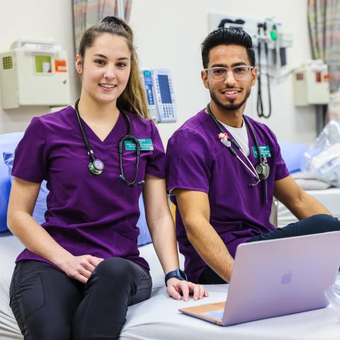 Two students in purple scrub shirts and black pants sit on an exam bed with a laptop.