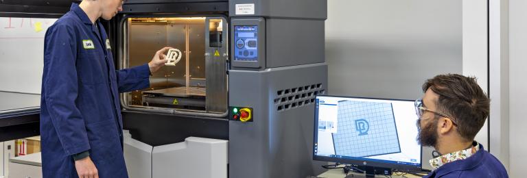 A student is pulling a 3D printed RDP logo out of a 3D printer while a faculty member watches