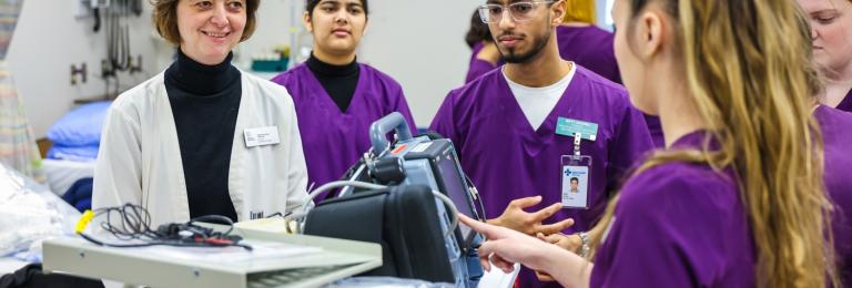 A group of nursing students in purple scrubs working on equipment while instructor watches