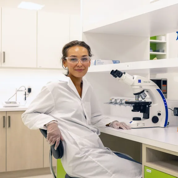 A researcher turns towards the camera, leaning their arm on the back of their chair, beside them is a microscope and various bottles.