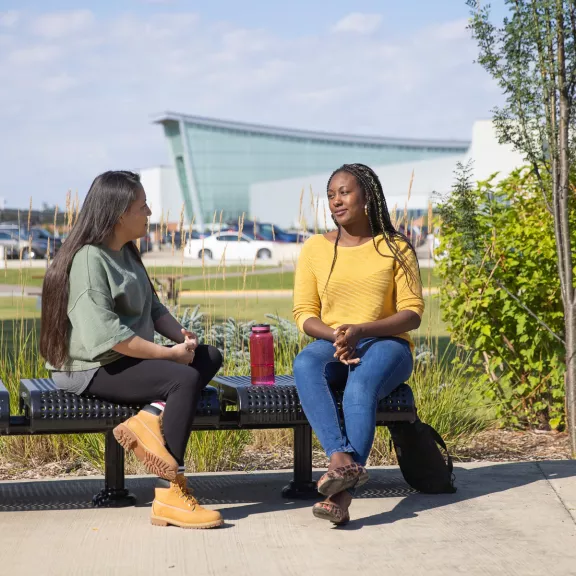 Two female students sit on a bench outside and chat