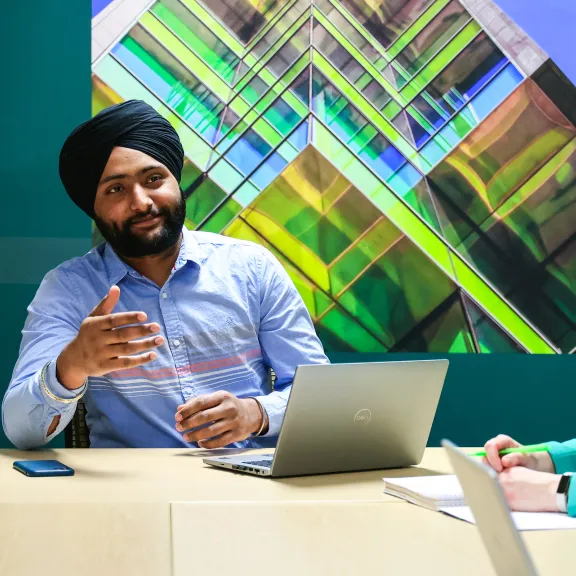 A group of professionals at a board table in a green room. On the wall hangs a large photo of a skyscraper with bright green segments dividing each floor. A man in a blue shirt and black headwrap smiles while talking to his coworkers whose faces are not shown to camera.