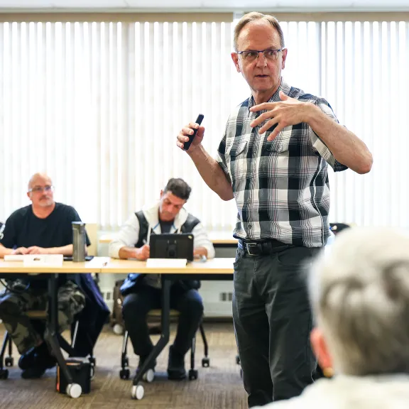 An instructor speaks to a class seated around him at tables against a bank of windows.