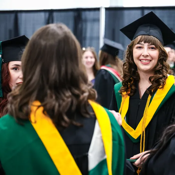 A group of graduates in gowns and caps gather, one smiles at the camera.