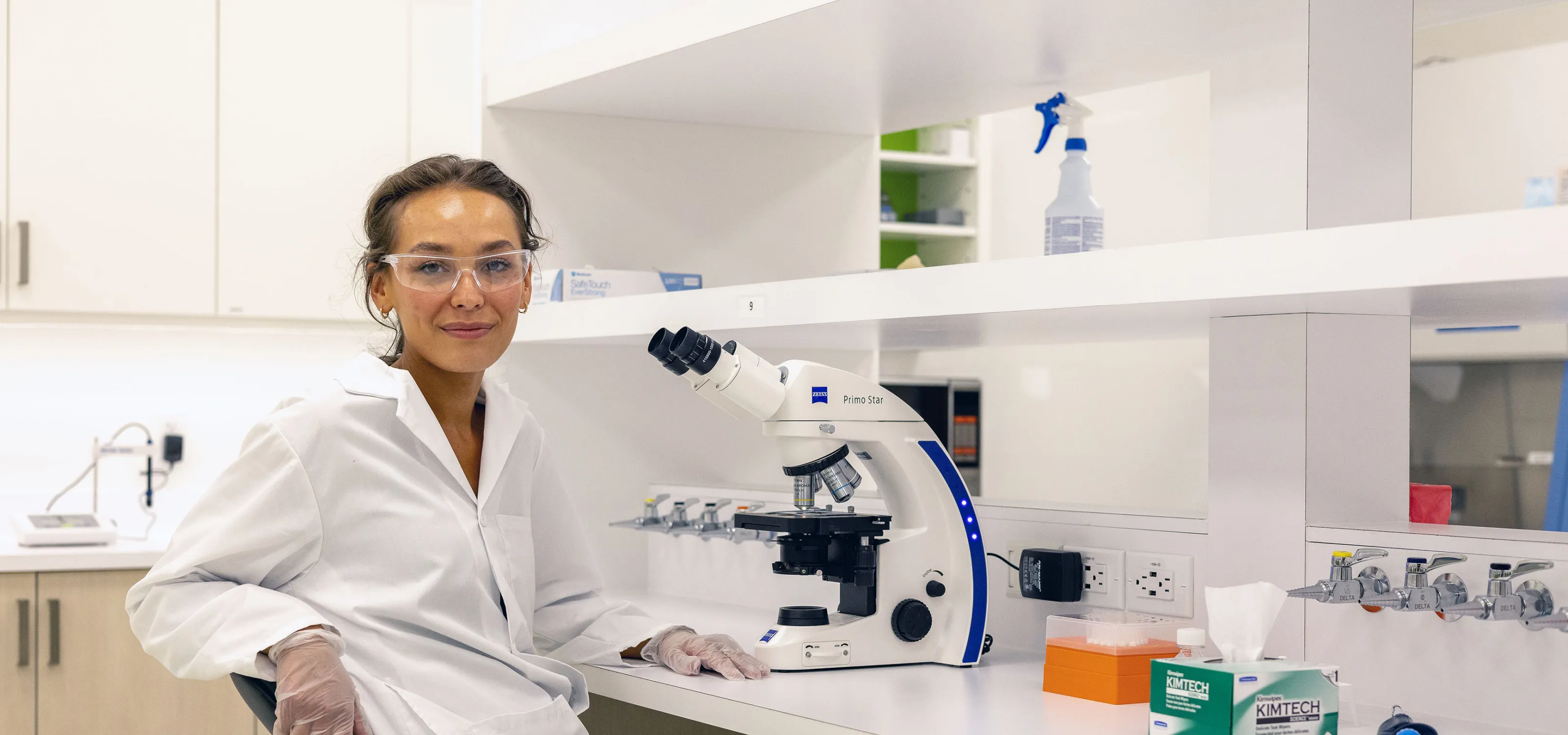 A researcher turns towards the camera, leaning their arm on the back of their chair, beside them is a microscope and various bottles.