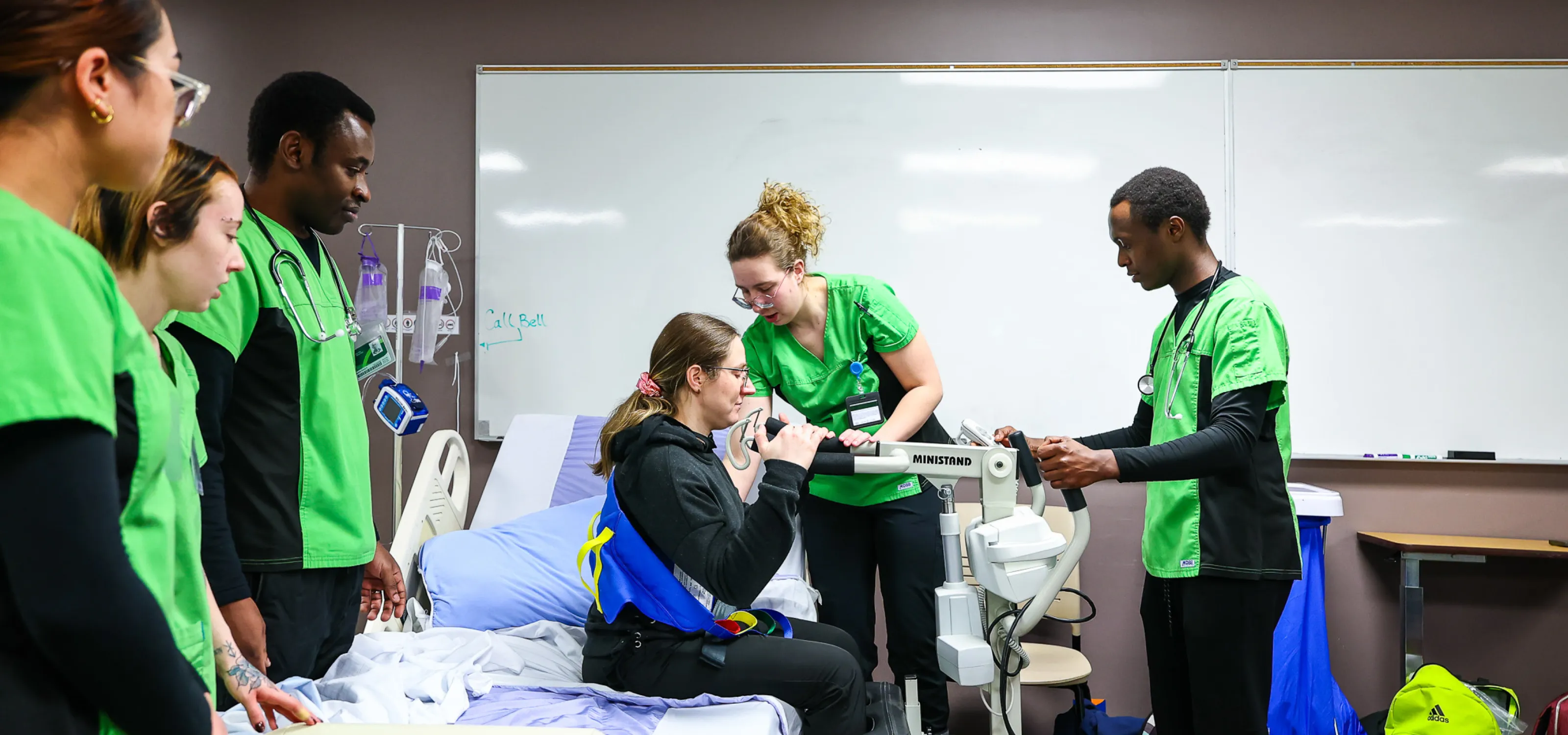 health care aide students, in lime green scrubs, help a patient stand up from a seated position using assistive device.