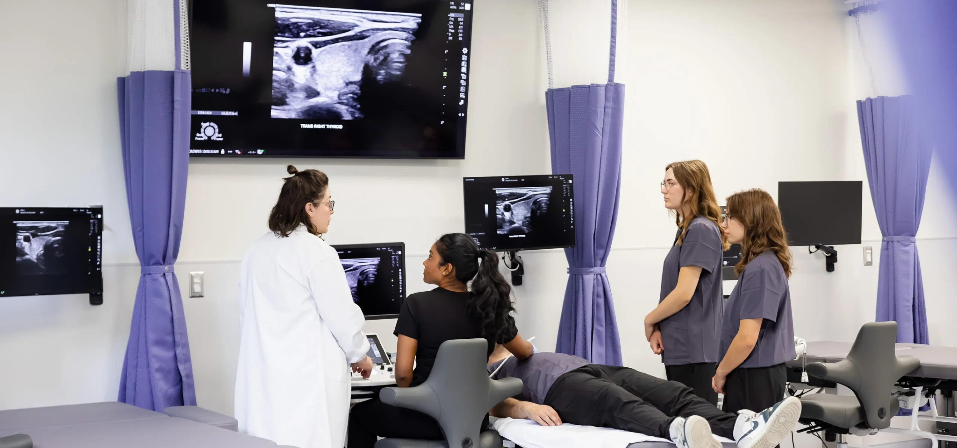 Students, some sitting standing and one laying on the bed, listen to an instructor while looking at an ultrasound image on various screens.